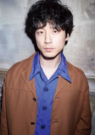 Portrait of a middle-aged Asian man with curly hair wearing a brown jacket over a blue shirt, standing against a textured background.
