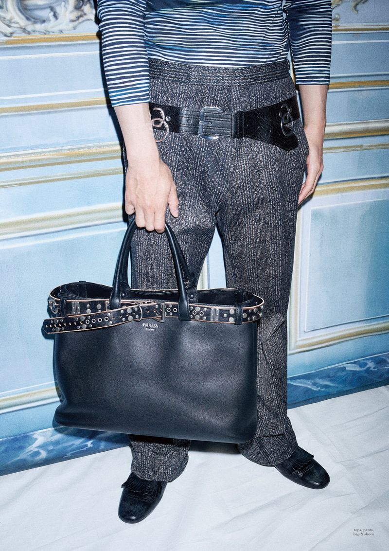 Fashion shot featuring a person holding a studded black leather Prada bag, wearing grey tweed pants, and a striped shirt against a blue wall.