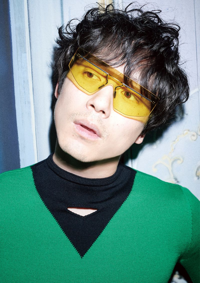 Stylish man with curly hair, wearing yellow sunglasses and a green sweater with a black collar, poses indoors against a patterned wall.