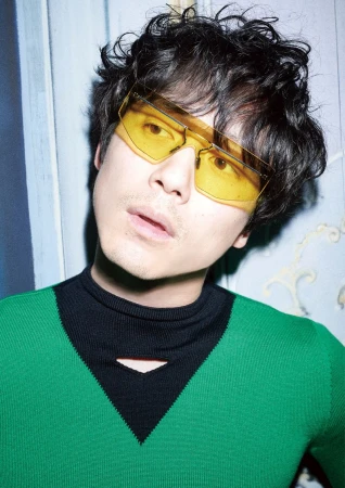 Stylish man with curly hair, wearing yellow sunglasses and a green sweater with a black collar, poses indoors against a patterned wall.