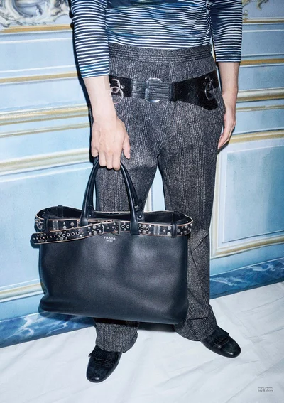Man wearing a striped top and grey tweed trousers holding a black leather tote bag against an ornate interior wall