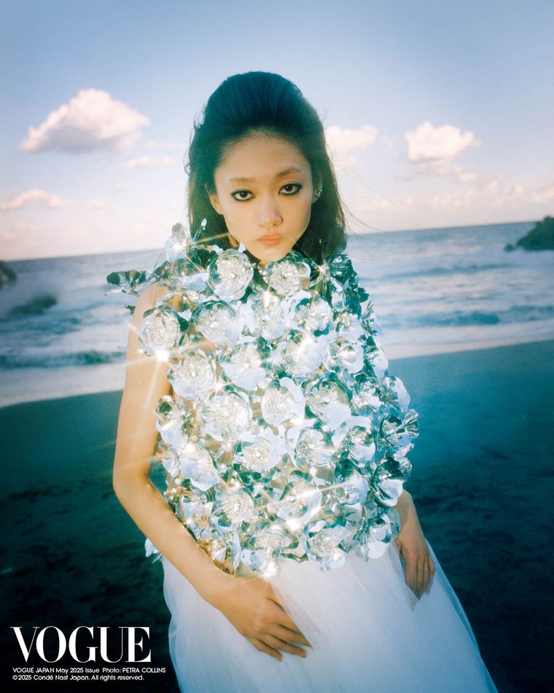 Young woman posing on a beach in a silver flower dress for a Vogue cover, featuring ocean, sky, and sand in the background.