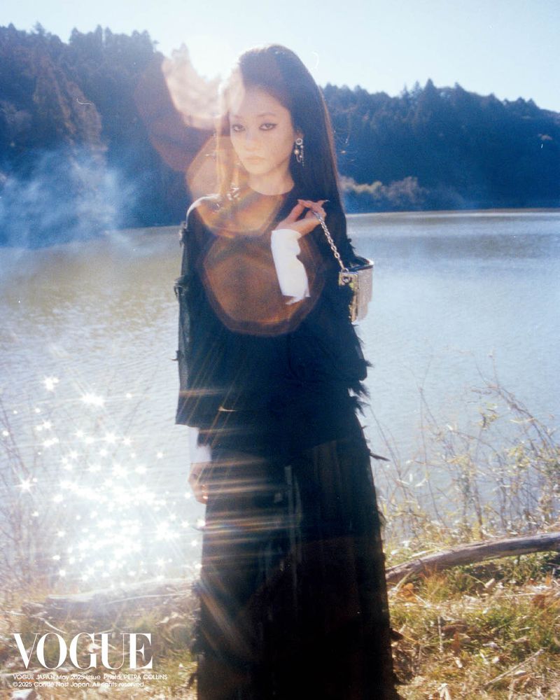 Young woman in a flowing black dress poses outdoors by a lake with mountains and trees under a bright sky, Vogue logo in the bottom left.