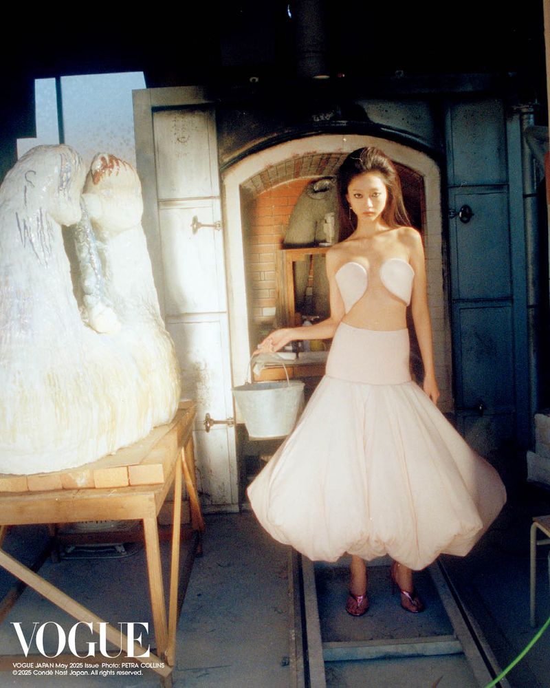 Fashion editorial shot of an Asian woman in a white dress holding a bucket in a rustic, industrial setting with large sculptures.