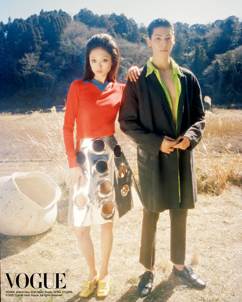 Two fashion models pose in a field; the woman wears a red top and silver skirt, while the man wears a green shirt and black coat.