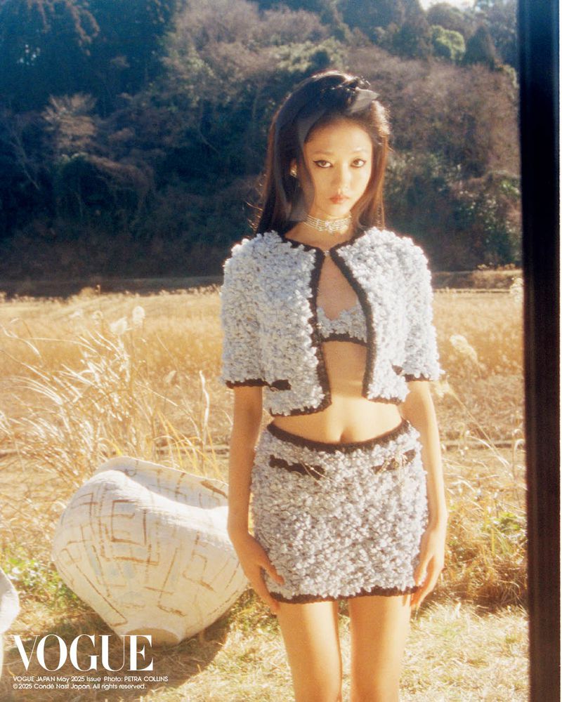 A woman in a tweed set poses in a field near an abstract sculpture under natural light for a Vogue Japan editorial fashion photograph.