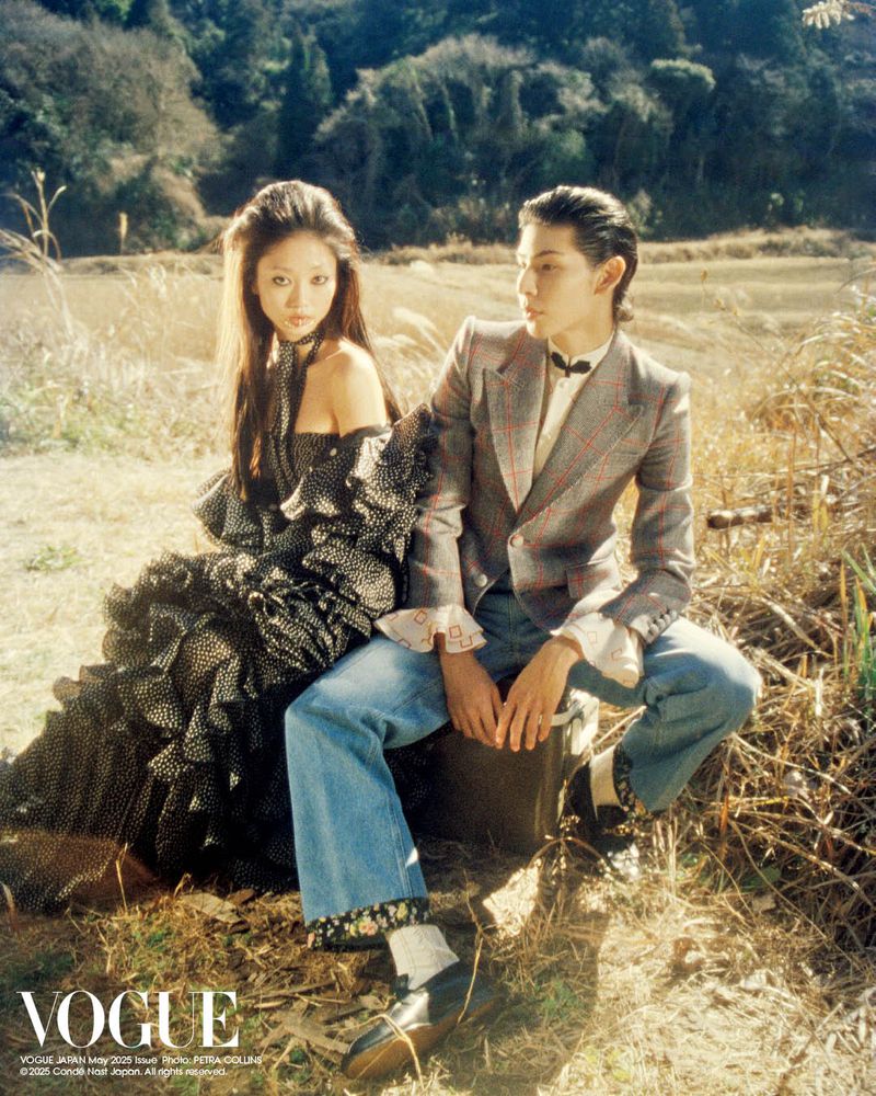 Asian couple in Vogue Japan photoshoot. The woman wears a black polka-dot dress, the man a gray suit and jeans, in a natural outdoor setting.
