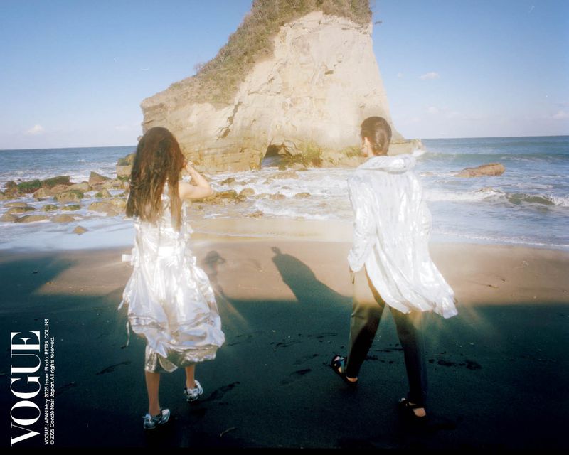 Two people walk on a black sand beach in front of a rock formation with an archway, on a sunny day, wearing fashionable clothes.