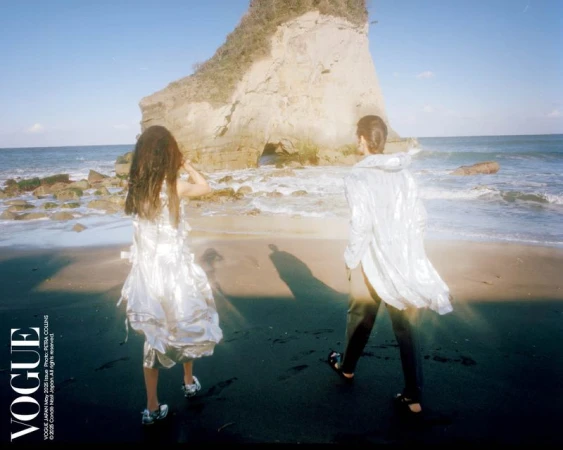 Two people walk on a black sand beach in front of a rock formation with an archway, on a sunny day, wearing fashionable clothes.