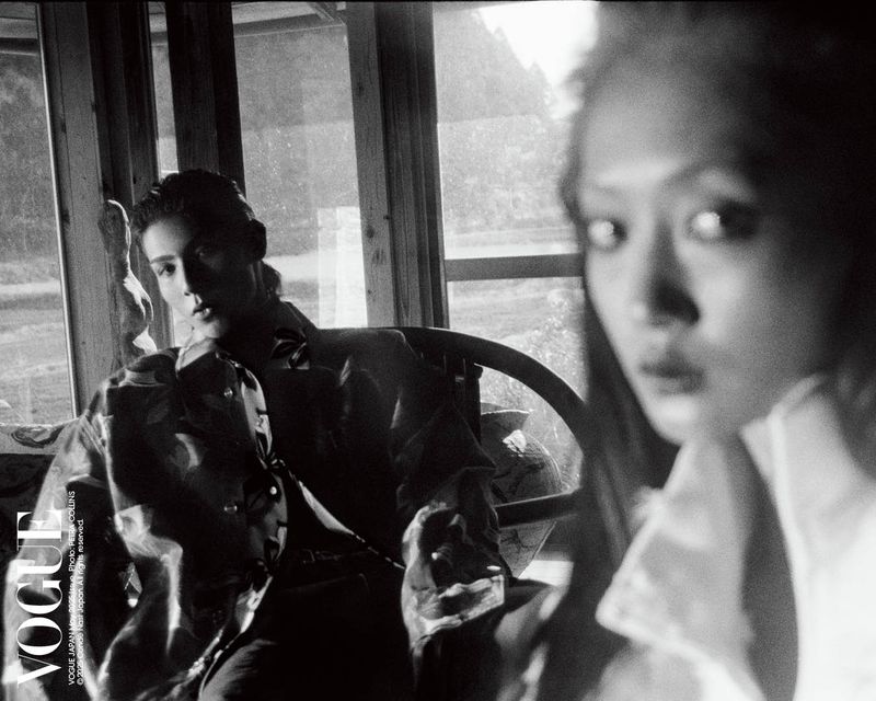 Black and white fashion shot with a model seated near a window and another woman in the foreground, features a Vogue logo.