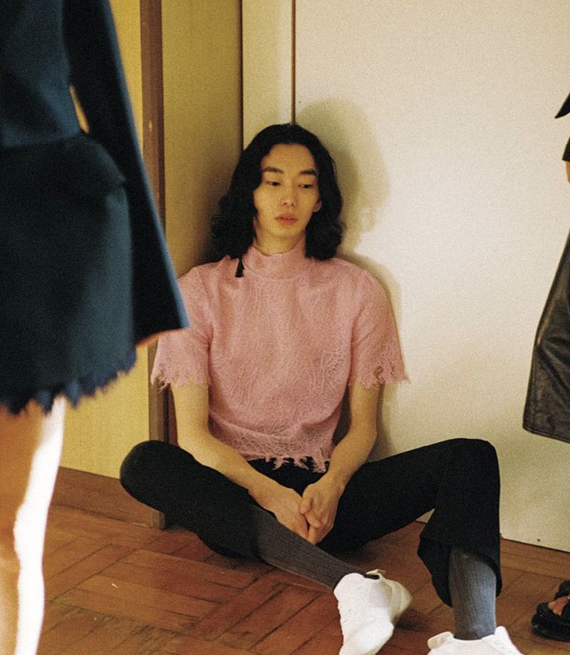 A person with long, dark hair sits on a wooden floor, wearing a pink shirt and black pants, with partial figures visible nearby.