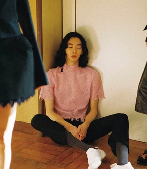 Model with long dark hair wearing a pink lace top sitting on a wooden floor in a hallway.