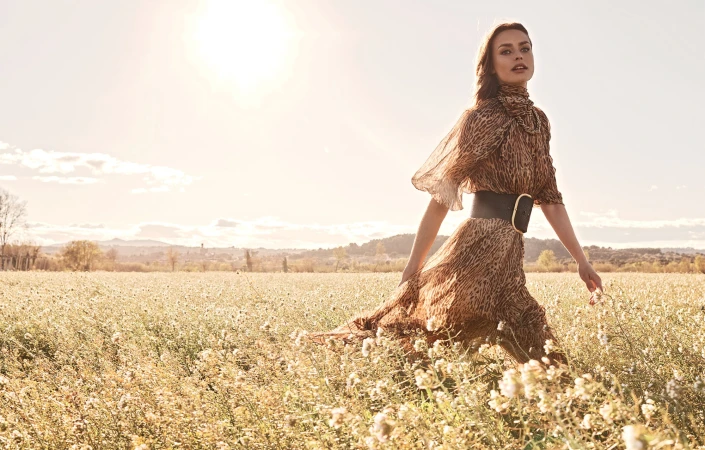 A woman walking through a sunny field of flowers wearing a patterned dress with a wide black belt.