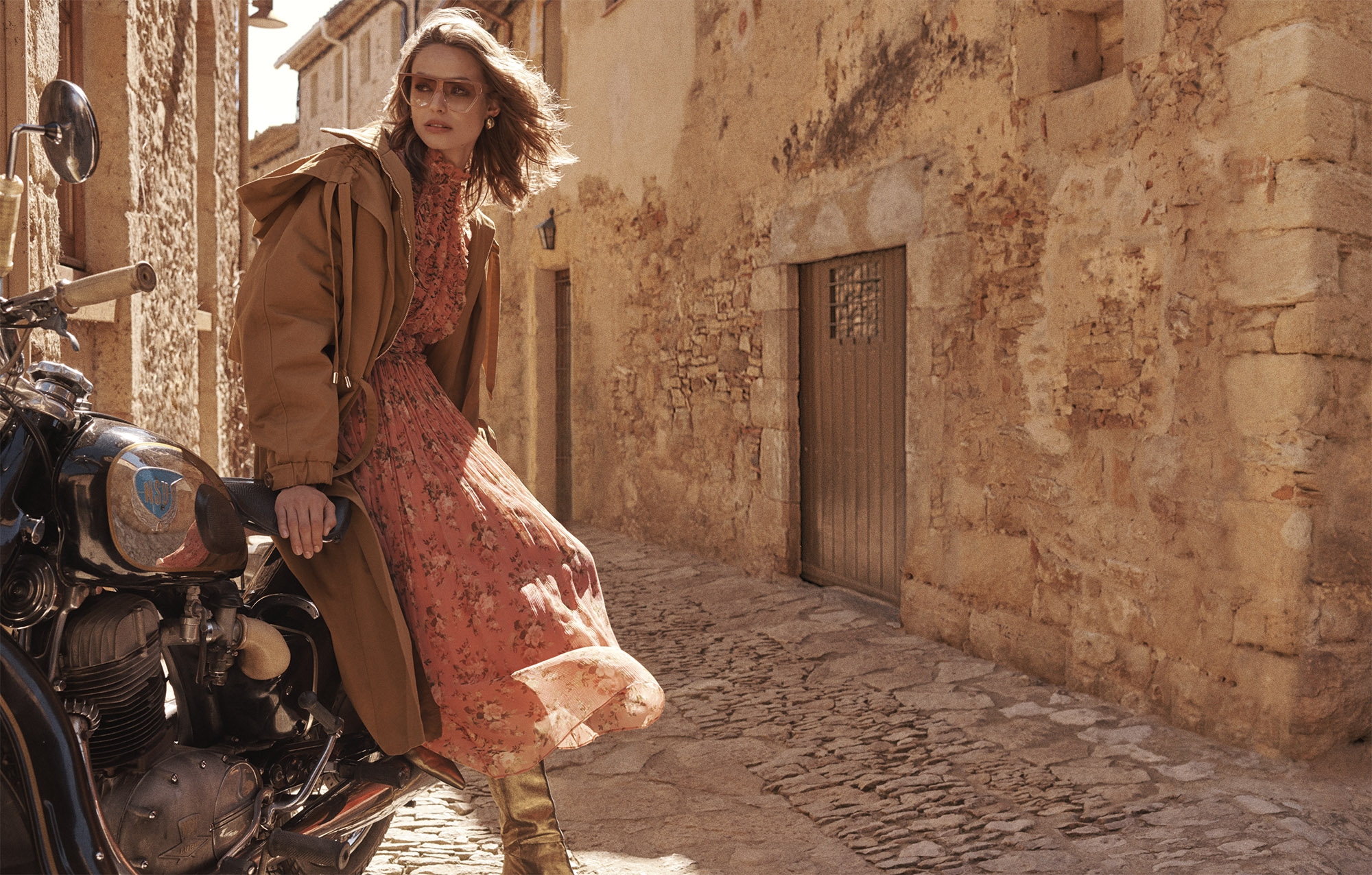 A fashionable adult female sits sideways on a vintage motorcycle in an outdoor urban setting, adorned in a long dress and brown coat.