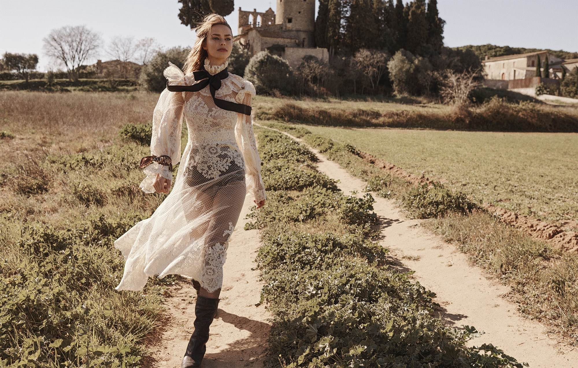 A young adult woman in a white lace dress and black boots walks on a dirt path through a field of green vegetation.