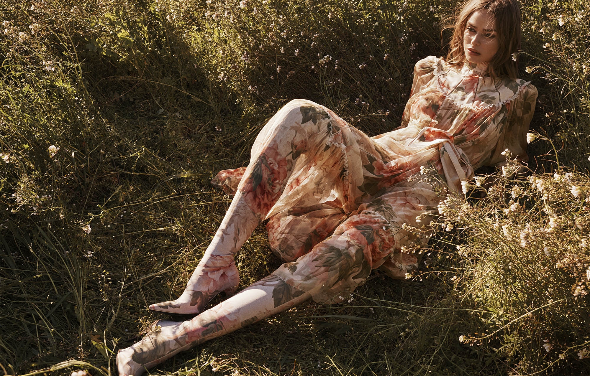 Woman in floral dress lies in grassy field with wildflowers in outdoor fashion shot, natural light, warm tones, soft focus.