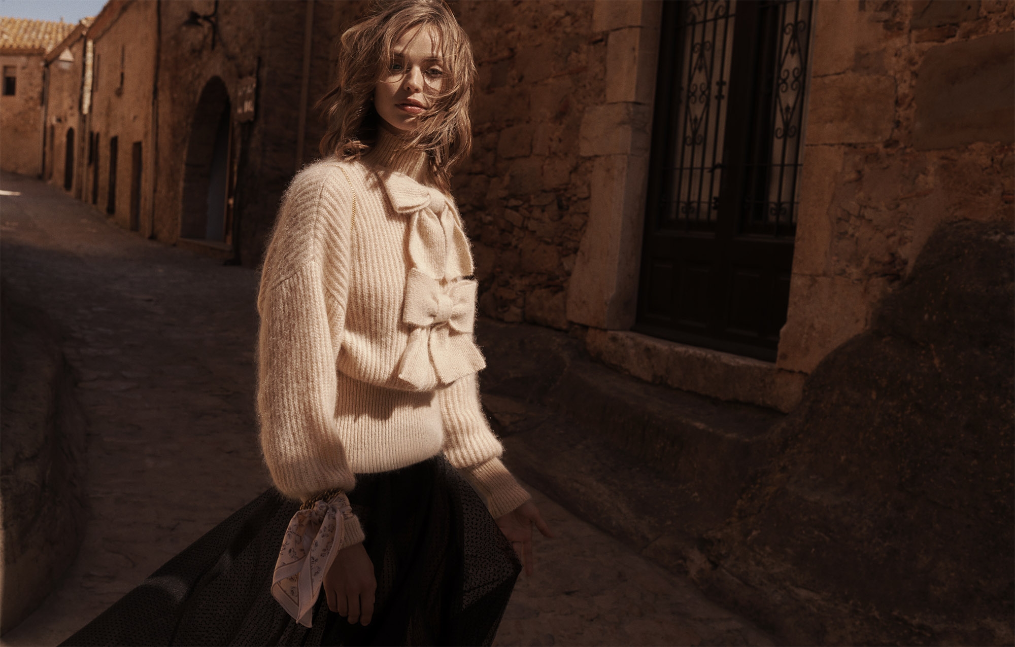 Fashion shot of a woman in a cream sweater and dark skirt walking down a stone street lined with buildings in warm, natural light.