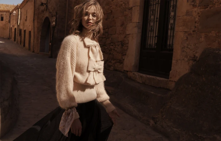 Fashion shot of a woman in a cream sweater and dark skirt walking down a stone street lined with buildings in warm, natural light.