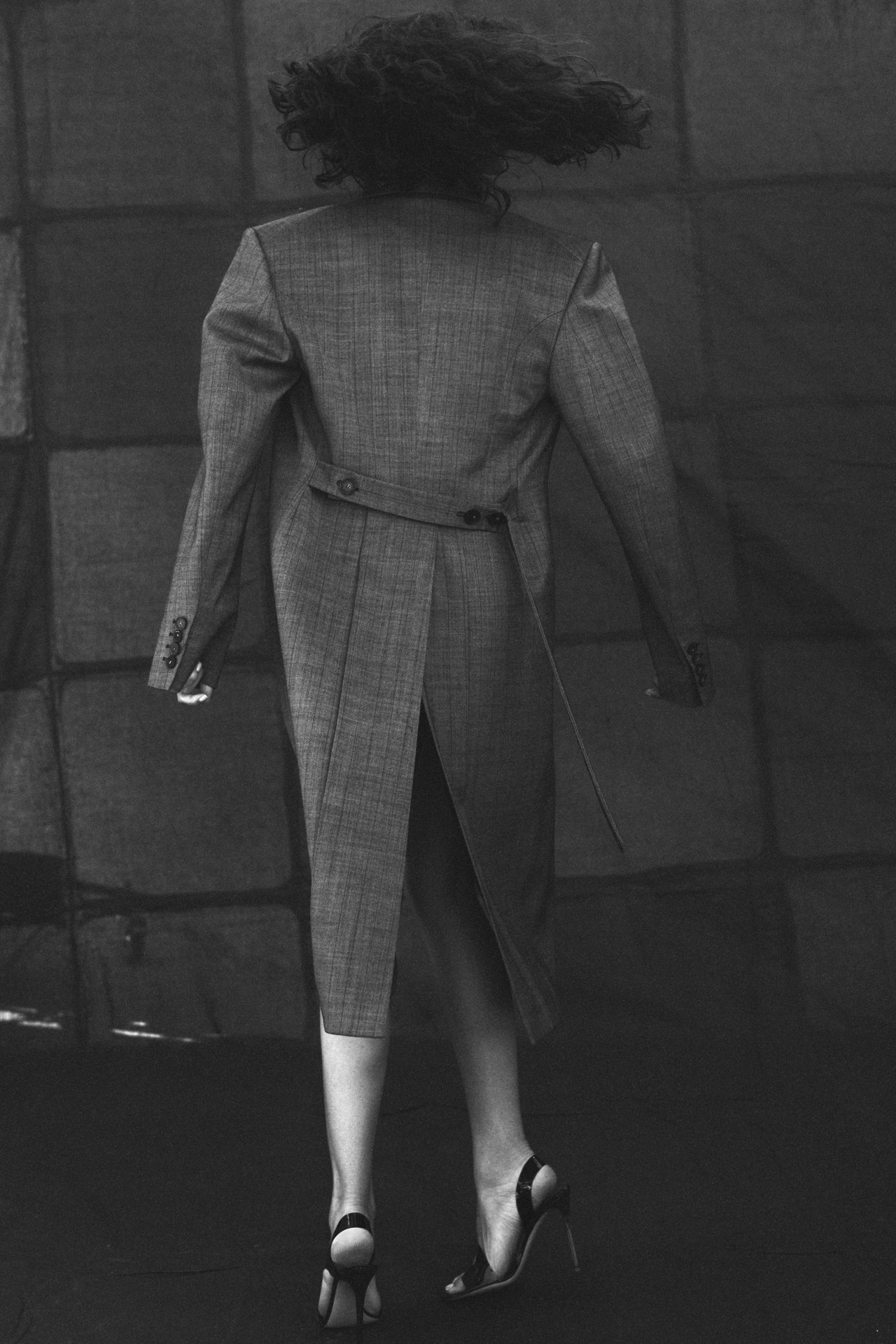 Back view of a woman with curly hair wearing a grey jacket dress and black high heels against a dark, geometric background.