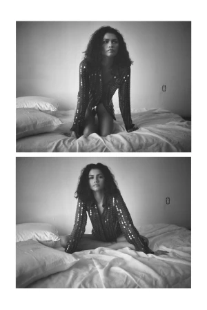 Zendaya kneeling and seated on a bed wearing a sequined long-sleeve shirt in a black and white editorial portrait photograph.