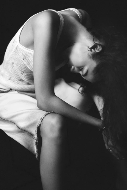 Monochrome portrait of a seated young adult female with long dark hair in a tank top and skirt, her head tilted down in a photo studio.