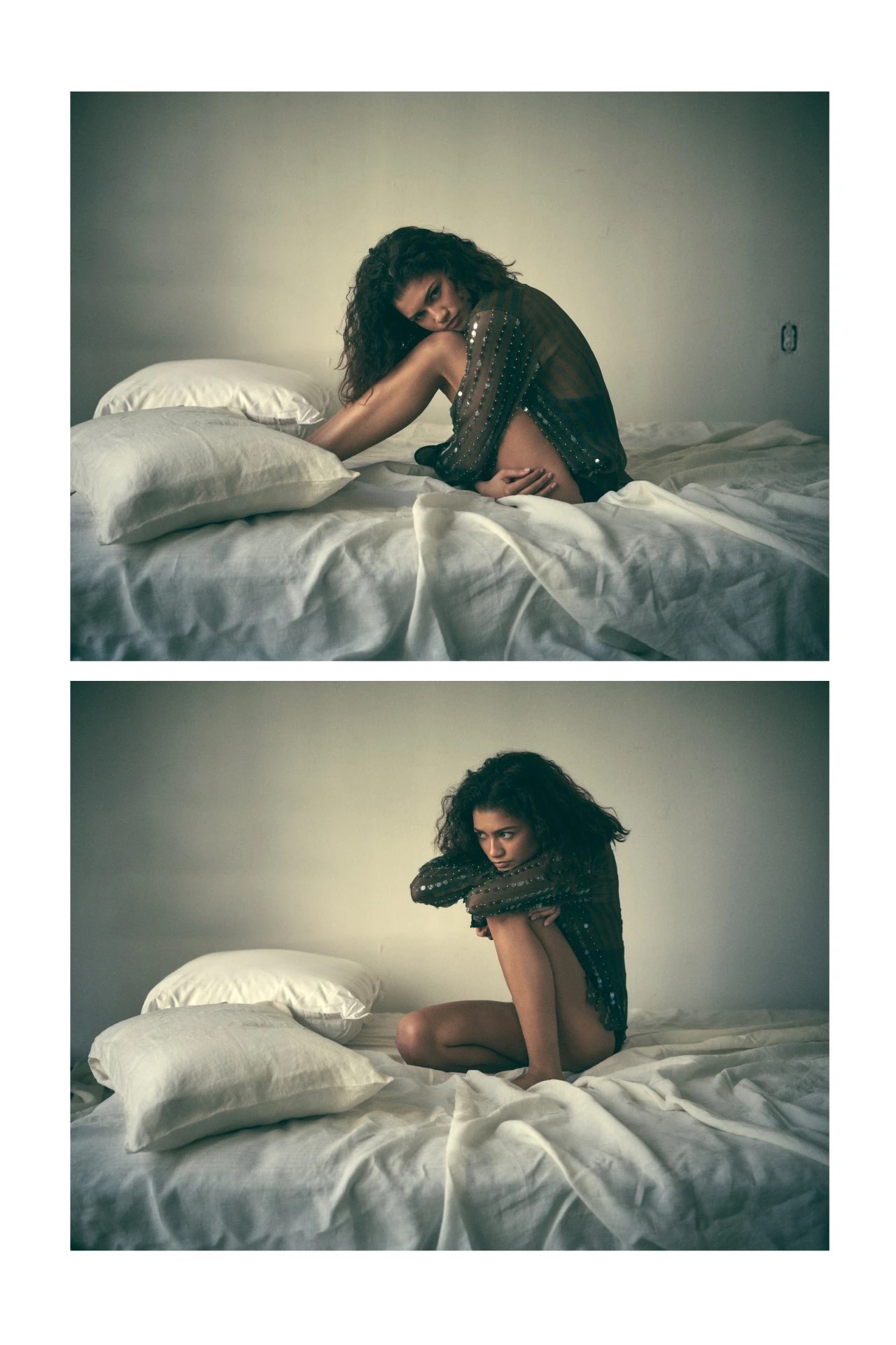 Brunette woman with curly hair poses on a bed in a translucent black long-sleeved top and underwear, with soft indoor lighting and white walls.