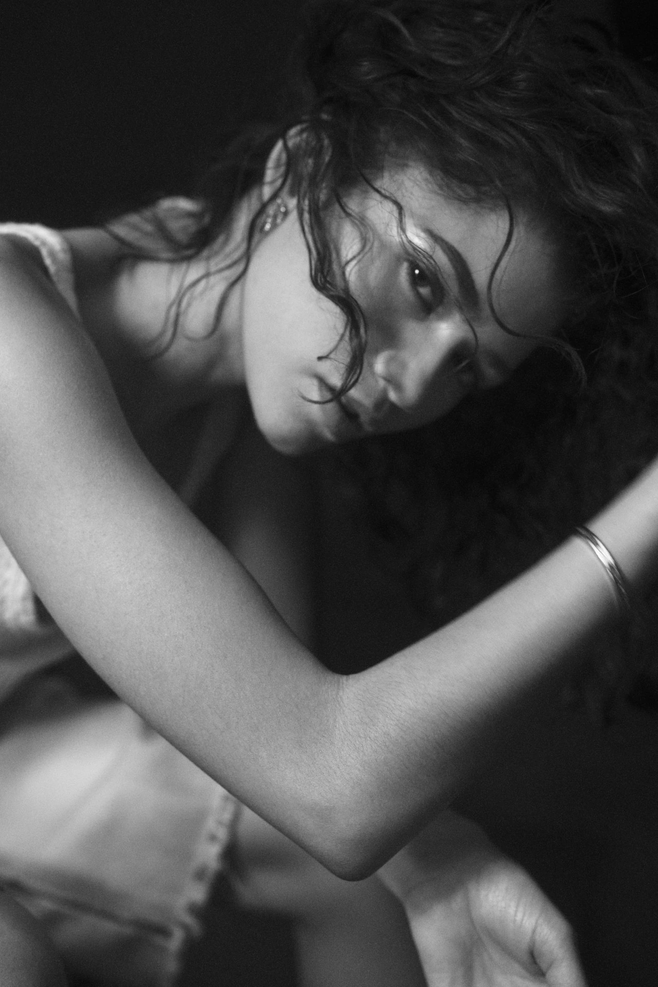 Black and white portrait of a young woman with curly hair and a contemplative expression, shot indoors with soft lighting on a dark background.