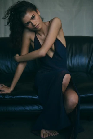 A young adult woman with curly hair sits on a black leather couch wearing a navy blue gown in a low-angle medium studio shot.
