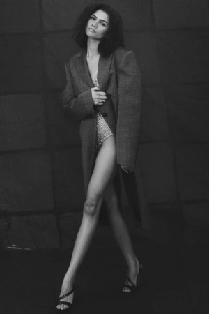 Zendaya poses in a grey coat and embellished bodysuit against a dark background in a studio portrait, showcasing a fashion-forward look.