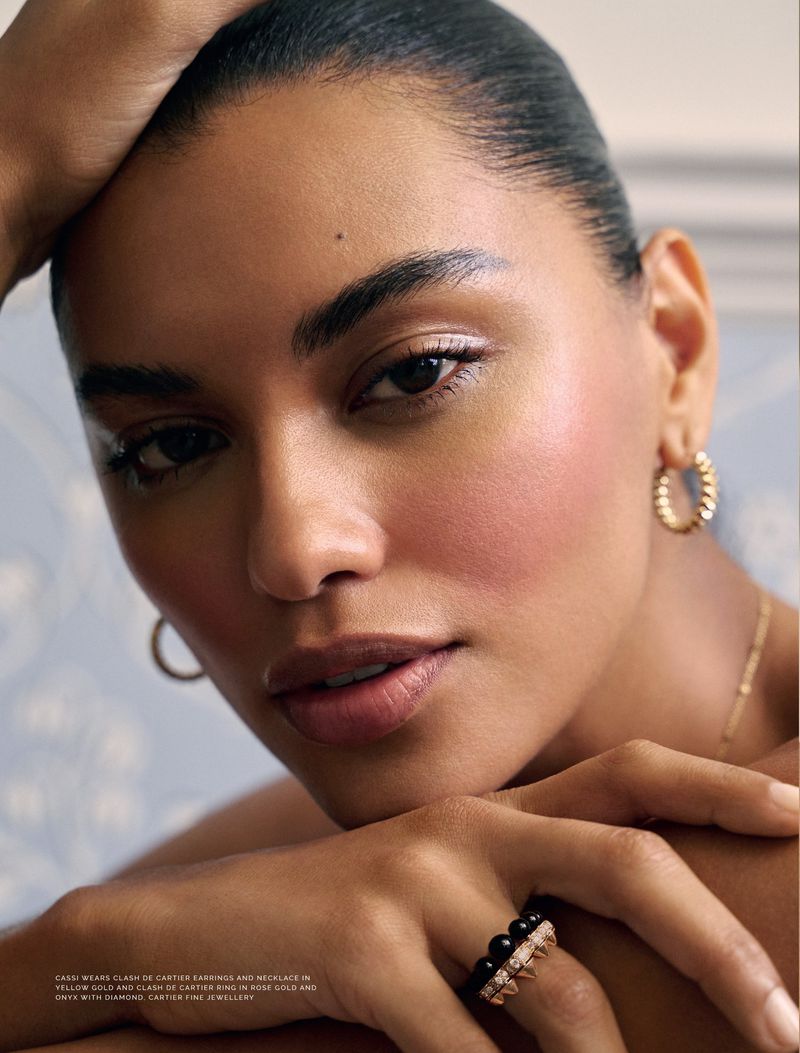 Close-up of a model wearing gold hoop earrings and a diamond ring, softly lit with natural light against a patterned backdrop.