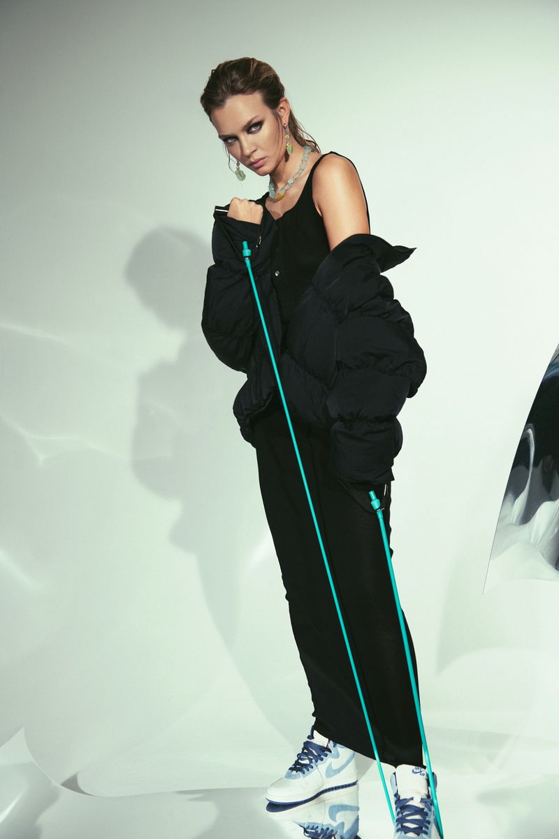 A woman in an all-black outfit poses in a studio. She wears a puffer jacket, sneakers, and jewelry, standing in front of a white background.