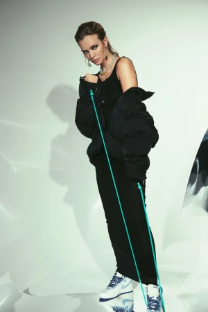 A woman in an all-black outfit poses in a studio. She wears a puffer jacket, sneakers, and jewelry, standing in front of a white background.