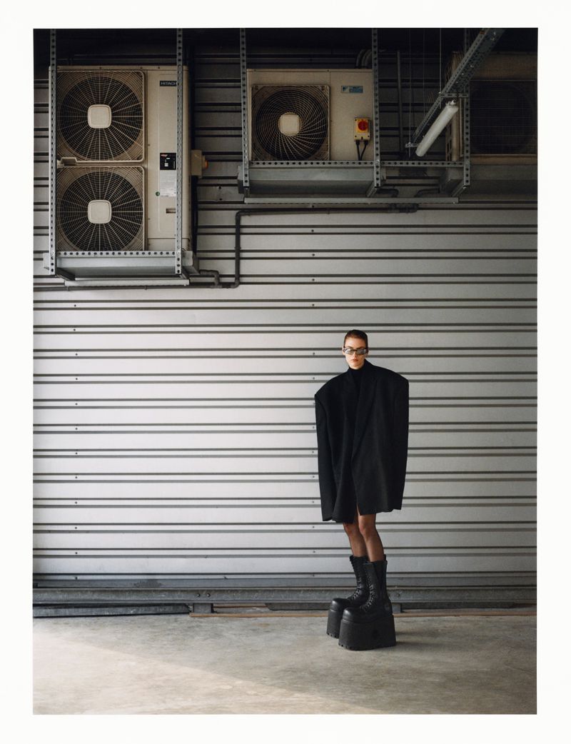 Fashion photography featuring a woman in a black outfit and platform boots standing against a corrugated metal wall with industrial elements.