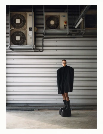 Fashion photography featuring a woman in a black outfit and platform boots standing against a corrugated metal wall with industrial elements.