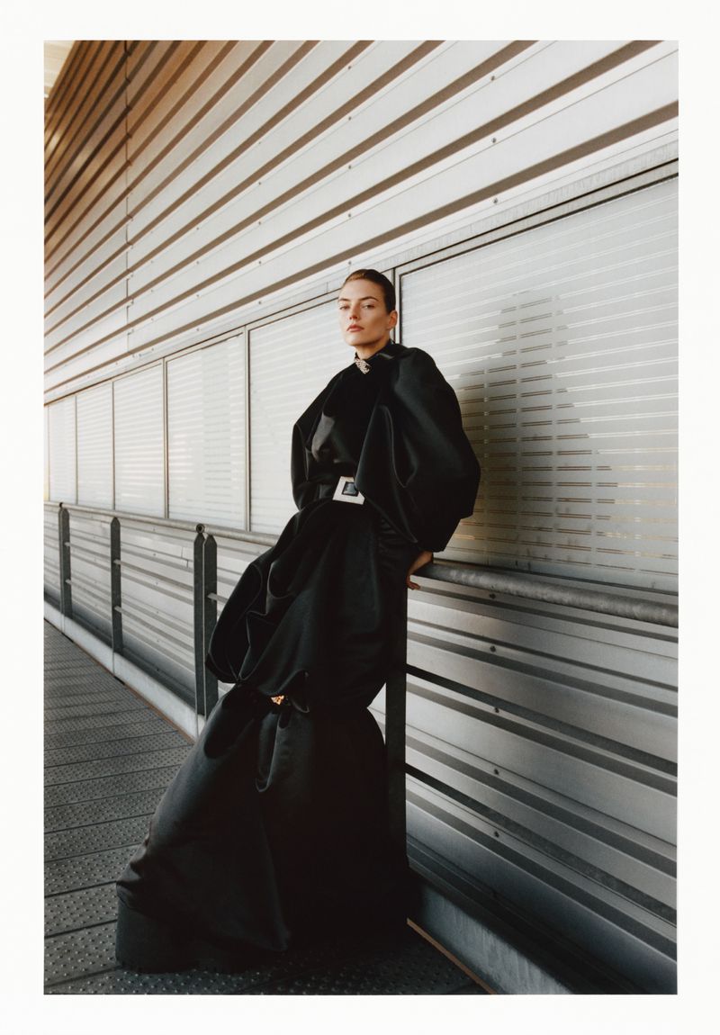Model in a black sculptural dress leaning against a metallic industrial wall for Exhibition Magazine Ti Amo project