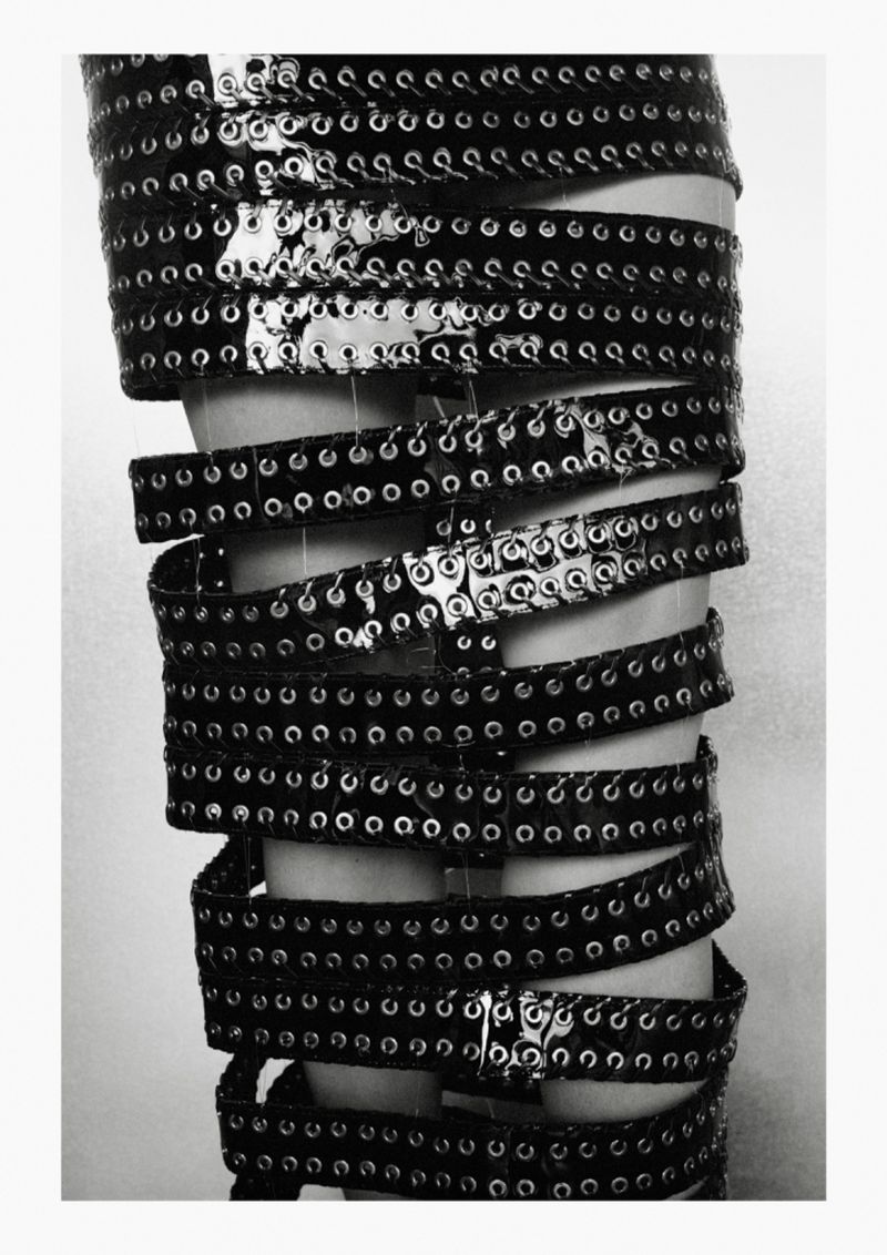 Closeup of a leg wrapped in multiple layers of shiny black grommeted belts against a plain light background in a fashion editorial.