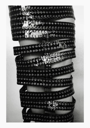 Closeup of a leg wrapped in multiple layers of shiny black grommeted belts against a plain light background in a fashion editorial.