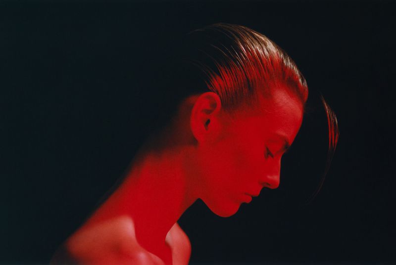 Profile portrait of a woman with slicked-back dark hair illuminated by vivid red light against a black background.