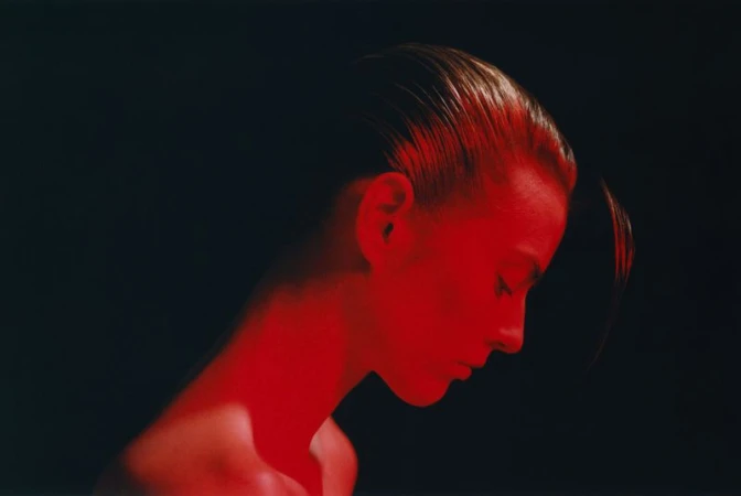 Profile portrait of a woman with slicked-back dark hair illuminated by vivid red light against a black background.