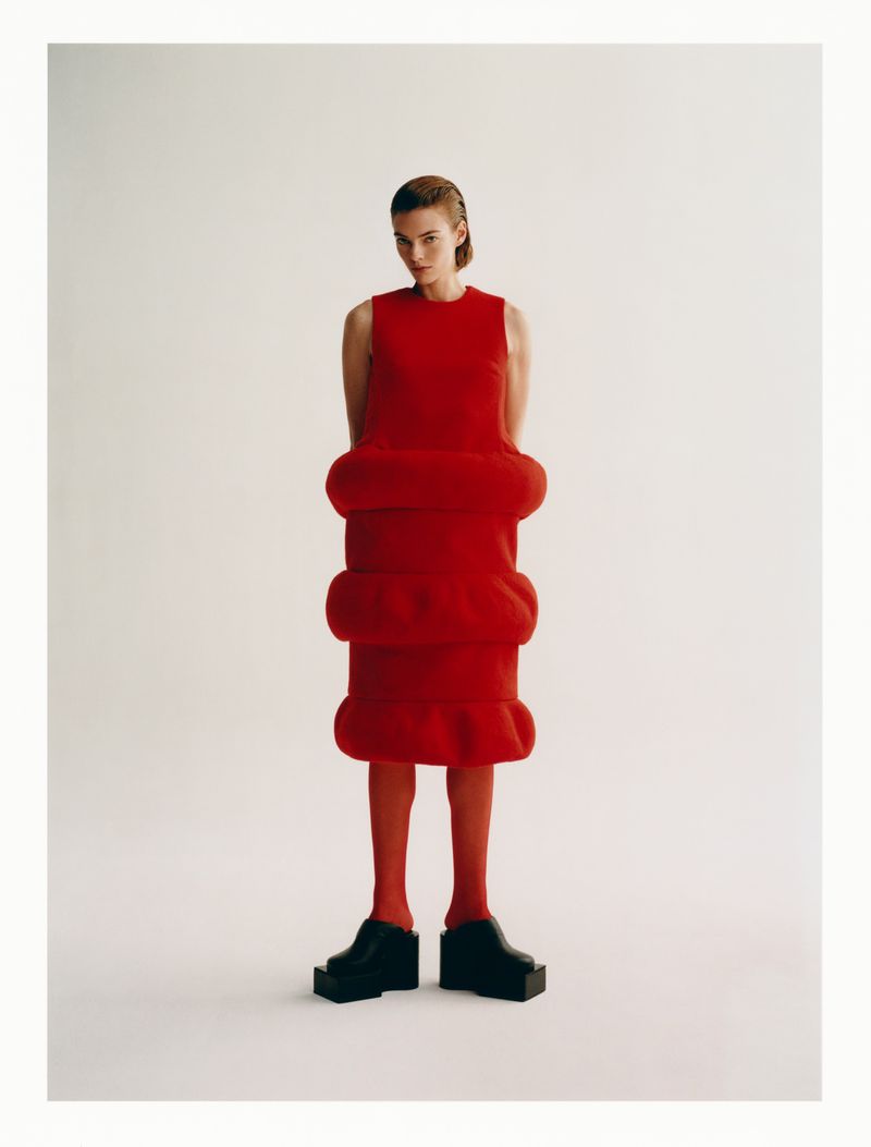 An adult woman in a red dress and red tights poses in a studio against a plain white background in a full-length portrait shot.