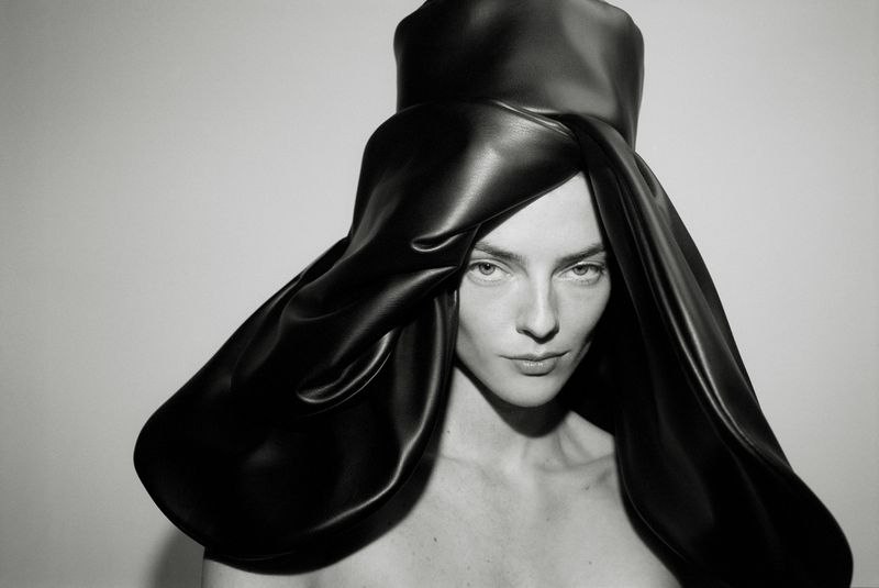 Monochrome portrait of a woman with light skin and dark hair wearing a dramatic black sculptural hat against a white background.