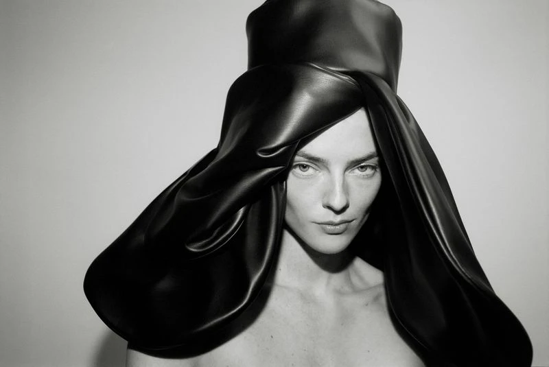 Monochrome portrait of a woman with light skin and dark hair wearing a dramatic black sculptural hat against a white background.