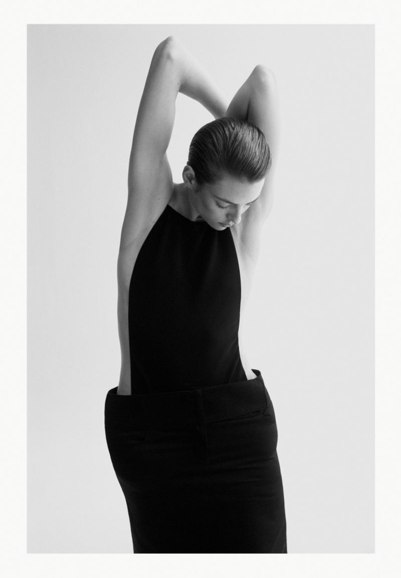 Monochromatic studio shot of a slender adult woman in a dark, elegant sleeveless dress, arms raised behind her head, looking down.