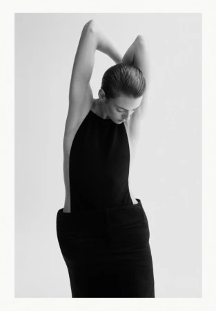 Monochromatic studio shot of a slender adult woman in a dark, elegant sleeveless dress, arms raised behind her head, looking down.