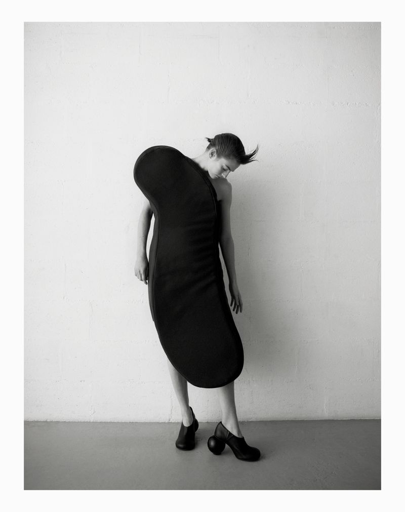 Monochrome fashion shot of a woman in an avant-garde dress and sculptural heeled shoes, posing against a textured white wall indoors.