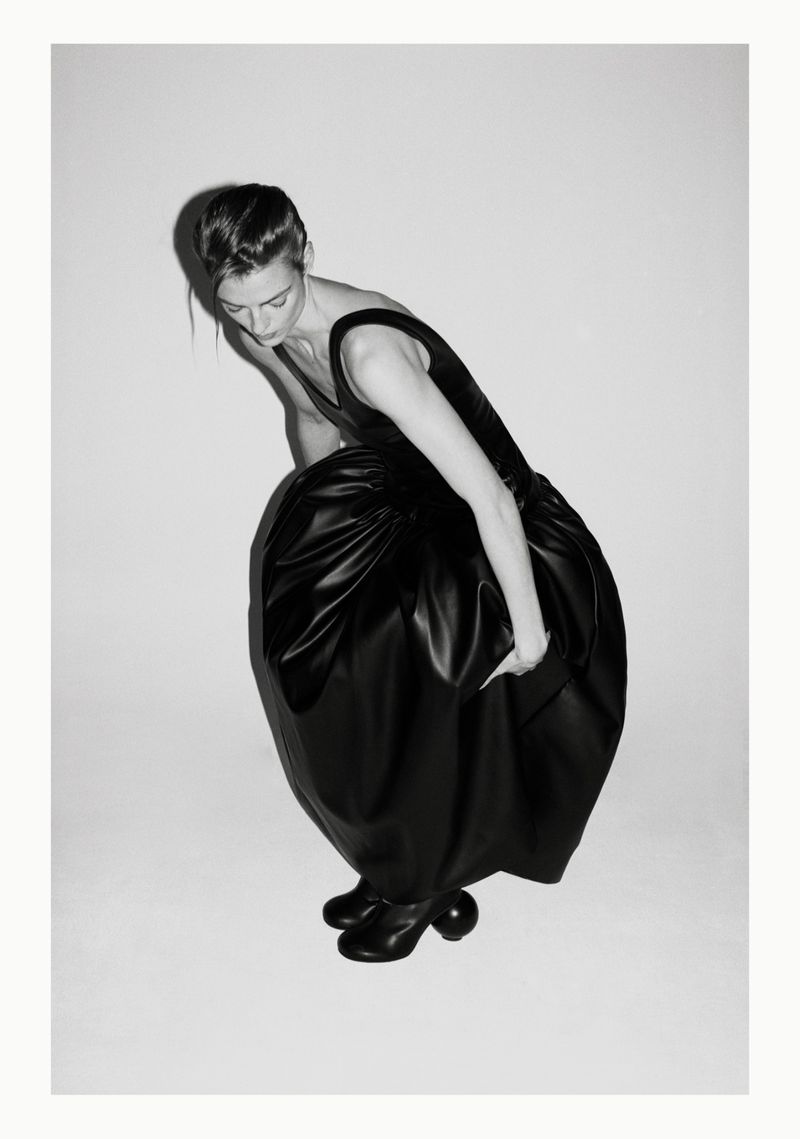 A woman in a black dress with a full skirt leans forward with her arm extended, shot in black and white against a plain backdrop.