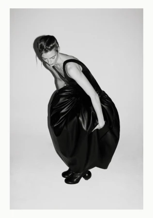 A woman in a black dress with a full skirt leans forward with her arm extended, shot in black and white against a plain backdrop.