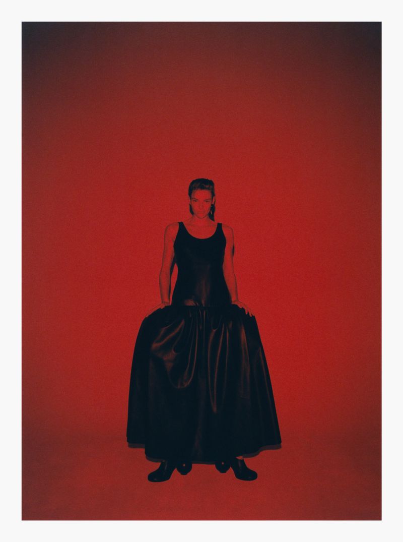 Model wearing a dark long dress standing against a vibrant red backdrop in a studio setting