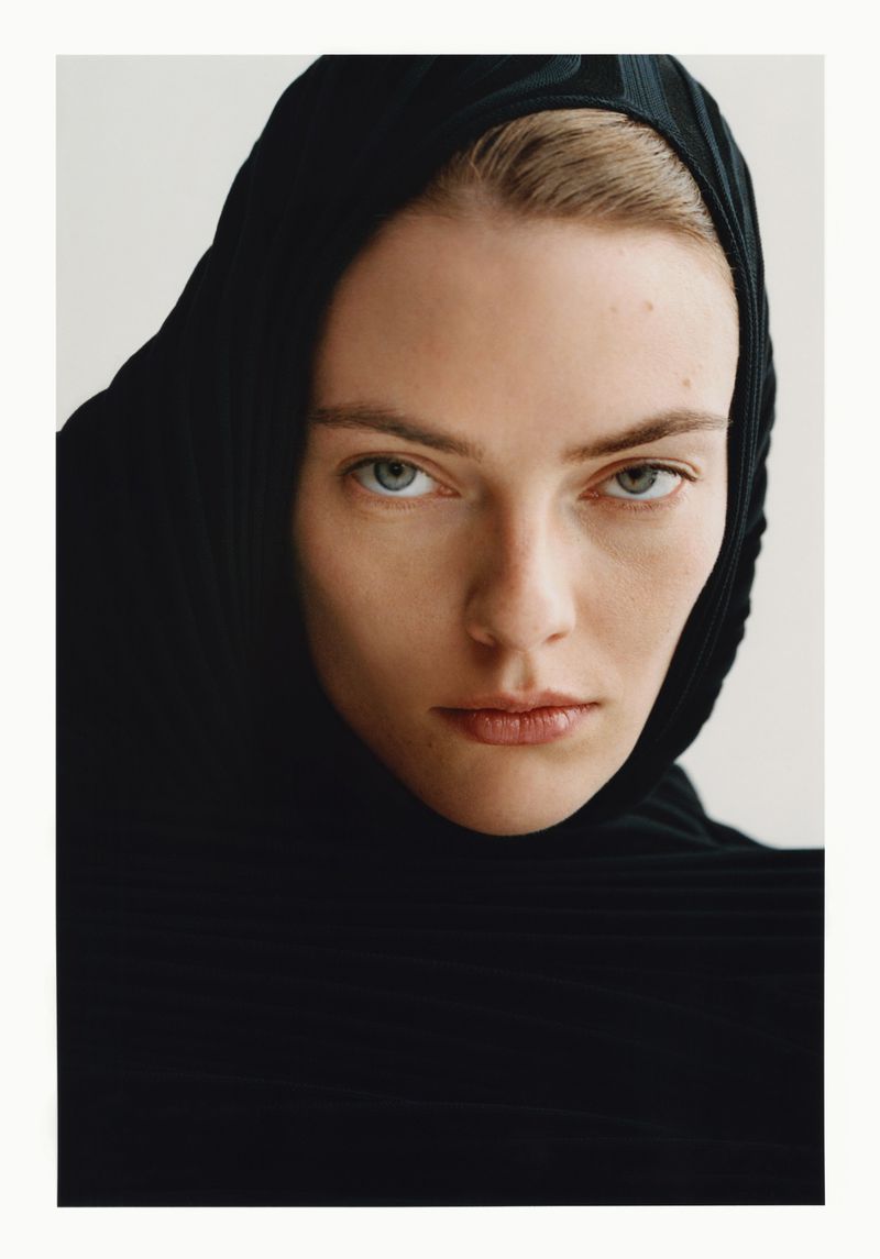Close-up portrait of a woman wearing a black hooded garment against a plain white background, showcasing a minimalist fashion aesthetic.