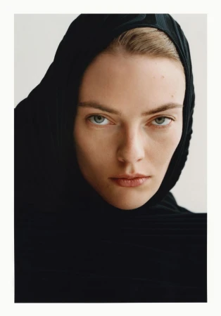 Close-up portrait of a woman wearing a black hooded garment against a plain white background, showcasing a minimalist fashion aesthetic.